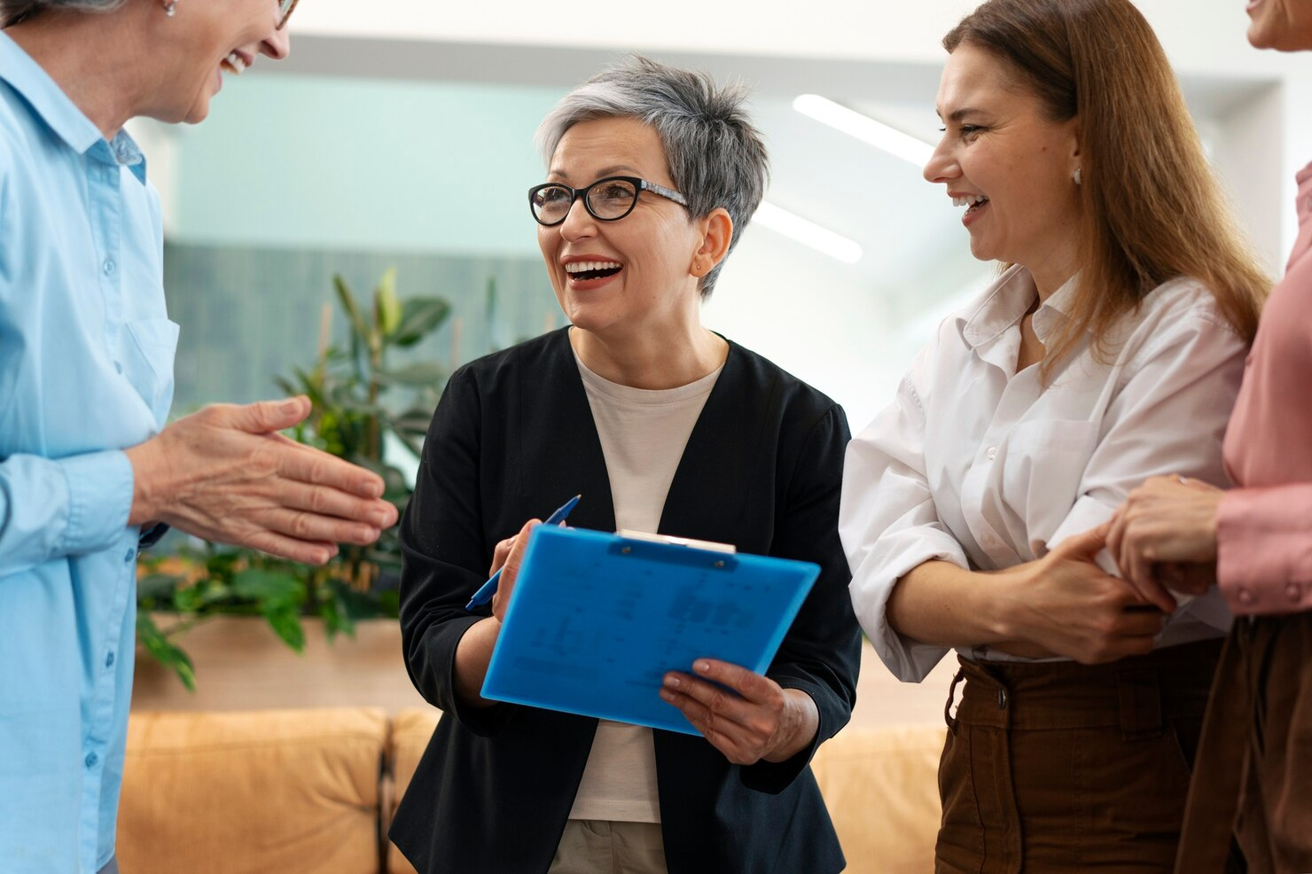 Three women gather around each other laughing while the middle woman holds a clip board and pen.